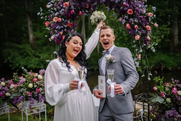 Valmiera, Letonya - 23 Mayıs 2025 - Şampanya kadehleri, buketler ve evlilik sertifikalarıyla çiçek kemeri altında neşeli bir gelin ve damat kutlaması.