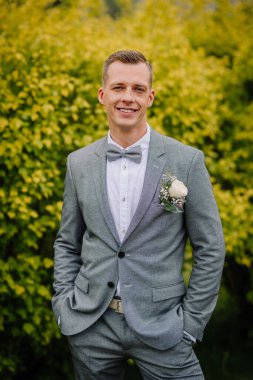 Gri takım elbiseli, elinde yaka çiçeğiyle gülümseyen damat, parlak yeşil ve sarı yapraklara karşı elleri ceplerinde poz veriyor..