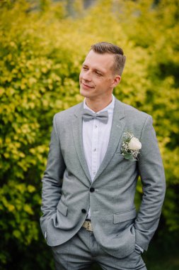 Yaka çiçekli açık gri bir takım elbise içinde damat, elleri ceplerinde, yumuşak bir gülümsemeyle yan yana bakan, canlı yeşil-sarı yaprakların önünde duran.