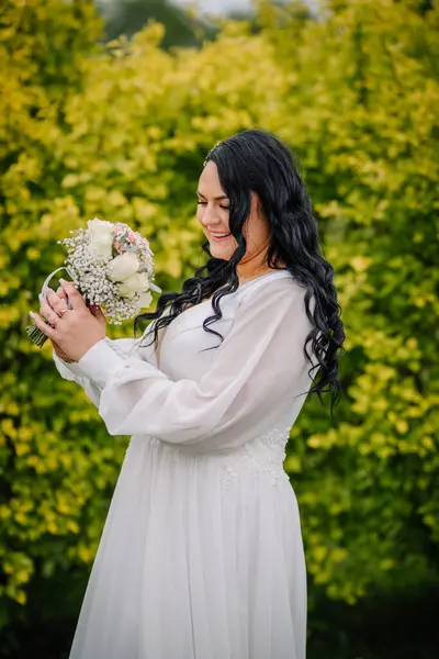 Beyaz elbiseli gelin, beyaz ve pembe çiçeklerden oluşan buketine bakarken nazikçe gülümsüyor. Parlak sarı-yeşil bir çitin önünde duruyor..