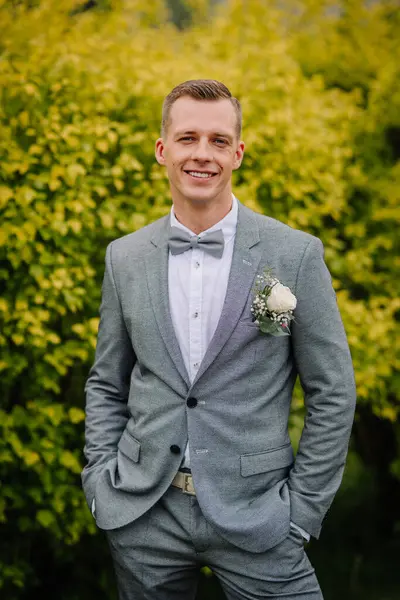 Gri takım elbiseli, elinde yaka çiçeğiyle gülümseyen damat, parlak yeşil ve sarı yapraklara karşı elleri ceplerinde poz veriyor..
