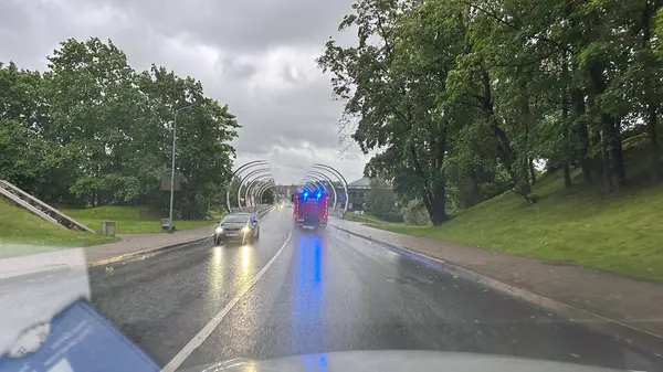 Valmiera, Letonya - 9 Haziran 2025 - Parlayan ışıklarla dolu itfaiye aracı yağmurlu bir günde arka planda yeşil ağaçlar ve modern kemer yapılarıyla ıslak bir şehir yolunda ilerliyor..