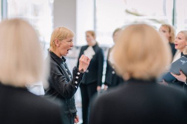 Valmiera, Letonya - 31 Mayıs 2025 - Bir kadın koro şefi, parlak bir odada resmi siyah kıyafetler giymiş şarkıcılar tarafından çevrelenmiş, anlamlı jestlerle vokal provasını yönetiyor..