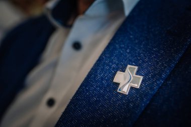 Valmiera, Letonya - 31 Mayıs 2025 - Resmi bir etkinlikte siyah düğmeli beyaz bir gömleğin üzerine giyilen, mavi bir haç rozetli mavi ceketin yakın çekimi.