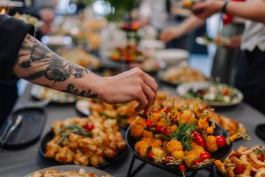 Valmiera, Letonya - 31 Mayıs 2025 - Bir sosyal ya da yemek organizasyonu sırasında çeşitli aperatiflerle dolu canlı bir büfe masasında renkli şişler için uzanan el dövmesi.