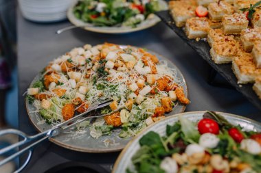 Kızarmış peynir ve kızarmış tavuk parçaları ile kaplı Sezar salatası tabağı diğer salata ve tatlıların yanına, açık büfe masasına konur..