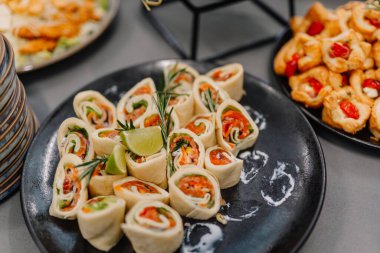 Açık büfedeki siyah servis tabağında limon ve biberiye ile süslenmiş sebze ve otlarla dolu düzgünce hazırlanmış tortilla dürümleri..