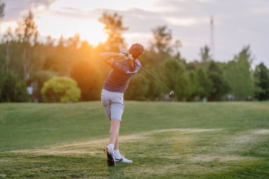 Valmiera, Letonya - 10 Haziran 2025 - Golfçü, yemyeşil ve ılık ışıklarla çevrili yemyeşil bir golf sahasında gün batımında bir salıncağı bitirir..