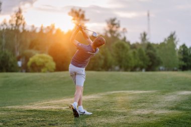 Valmiera, Letonya - 10 Haziran 2025 - Golfçü, yemyeşil ve ılık ışıklarla çevrili yemyeşil bir golf sahasında gün batımında bir salıncağı bitirir..