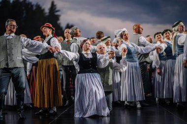 Valmiera, Letonya - 7 Haziran 2025 - Geleneksel Letonya kıyafetleriyle büyük bir grup halk dansçısı GAUJA 80 kutlamaları sırasında dramatik gökyüzü altında enerjik bir performans sergiliyorlar..