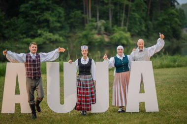 Valmiera, Letonya - 7 Haziran 2025 - Geleneksel Letonya kostümlü dört kişi, GAUJA 80 harfli çimlerin üzerindeki GAUJA 80 harfinin yanında gülümseyerek poz veriyor..