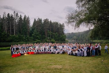 Valmiera, Letonya - 7 Haziran 2025 - GAUJA 80 'de renkli giyinmiş geleneksel Letonyalı halk dansçılarının toplu fotoğrafı, orman ve nehir zeminli yemyeşil bir çayırda poz verdiler..