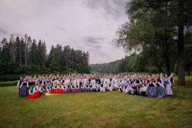 Valmiera, Letonya - 7 Haziran 2025 - GAUJA 80 'de renkli giyinmiş geleneksel Letonyalı halk dansçılarının toplu fotoğrafı, orman ve nehir zeminli yemyeşil bir çayırda poz verdiler..