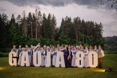 Valmiera, Letonya - 7 Haziran 2025 - Geleneksel Letonyalı halk dansçıları büyük GAUJA 80 harfli orman arkaplan manzaralı bir çayırda poz veriyorlar. Yıldönümü kutlaması
