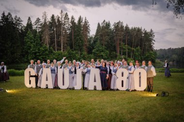 Valmiera, Letonya - 7 Haziran 2025 - Geleneksel Letonyalı halk dansçıları büyük GAUJA 80 harfli orman arkaplan manzaralı bir çayırda poz veriyorlar. Yıldönümü kutlaması