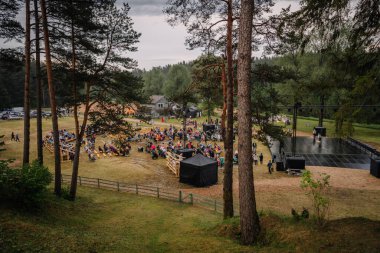 Valmiera, Letonya - 7 Haziran 2025 - GAUJA 80 açık hava kutlamaları, kalabalık bir göl ve sahne kurulumu, ağaçlar ve bulutlu gökyüzünün altında ahşap çitlerle çevrili.