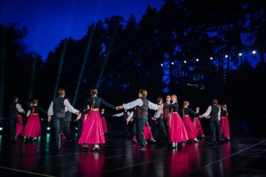 Valmiera, Letonya - 7 Haziran 2025 - GAUJA 80 'deki geleneksel Letonya halk dansları kolajı, renkli kostümlü dansçıları gece aydınlatmalı bir sahnede sergiliyorlar..