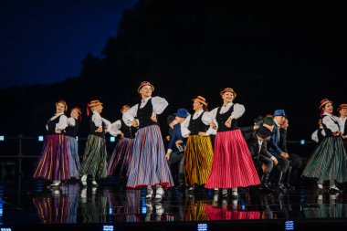 Valmiera, Letonya - 7 Haziran 2025 - GAUJA 80 'deki geleneksel Letonya halk dansları kolajı, renkli kostümlü dansçıları gece aydınlatmalı bir sahnede sergiliyorlar..