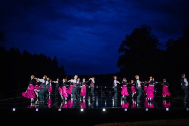 Valmiera, Letonya - 7 Haziran 2025 - GAUJA 80 'deki geleneksel Letonya halk dansları kolajı, renkli kostümlü dansçıları gece aydınlatmalı bir sahnede sergiliyorlar..