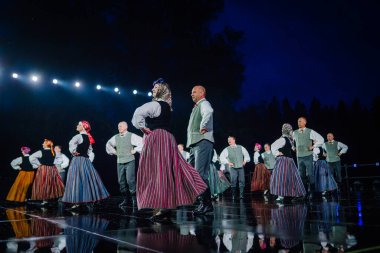 Valmiera, Letonya - 7 Haziran 2025 - GAUJA 80 'deki geleneksel Letonya halk dansları kolajı, renkli kostümlü dansçıları gece aydınlatmalı bir sahnede sergiliyorlar..