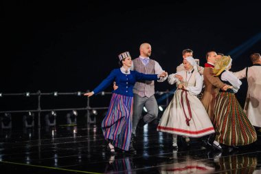 Valmiera, Letonya - 7 Haziran 2025 - GAUJA 80 'deki geleneksel Letonya halk dansları kolajı, renkli kostümlü dansçıları gece aydınlatmalı bir sahnede sergiliyorlar..