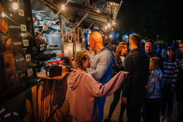 Cesis, Letonya 20 Haziran 2025 - Bir gece etkinliği sırasında insanlar canlı bir sokak yemek kamyonunda kuyruk ve etkileşim içinde, sıcak ışıklandırma, menü tahtaları ve renkli ambalaj görünür.