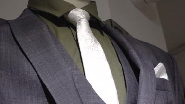 Gri kareli ceket, zeytin gömlek, beyaz desenli kravat ve vitrindeki cep mendili eşliğinde şık bir erkek takımının yakın çekimi..