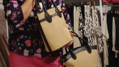 Valmiera, Letonya - 15 Haziran 2025 - Çiçekli bluzlu ve pembe etekli bir kadın bir moda mağazasında siyah saplı kamış bir el çantası tutuyor, modern monokrom kıyafetlerle çevrili.