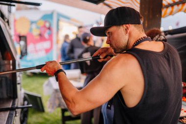 Liepa, Letonya - 27 Haziran 2025 - Siyah kolsuz tişörtlü ve şapkalı bir adam arka planda insanlar ve tezgahlar olan bir açık hava yemek kamyoneti festivalinde ahşap fırında yemek pişirmek için pizza kabuğu kullanıyor..