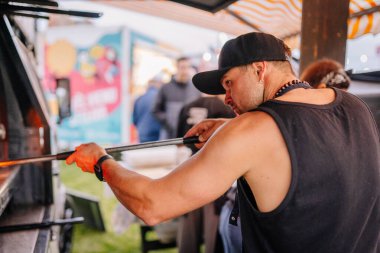 Liepa, Letonya - 27 Haziran 2025 - Siyah kolsuz tişörtlü ve şapkalı bir adam arka planda insanlar ve tezgahlar olan bir açık hava yemek kamyoneti festivalinde ahşap fırında yemek pişirmek için pizza kabuğu kullanıyor..