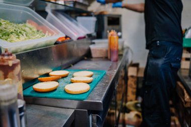 Liepa, Letonya - 27 Haziran 2025 - Burger çörekleri ticari bir mutfakta yeşil bir kesme tahtasına konur ve arka planda soslar ve sebzelerin yanında malzemeler hazırlayan bir işçi bulunur..