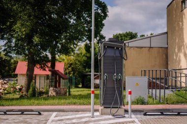 Strenci, Letonya - 21 Haziran 2025 - Üç etiketli elektrikli araç şarj istasyonu (A, B, C) bir binanın yanındaki otoparka yerleştirildi ve gün ışığı altında yeşillik.