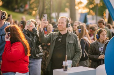 Cesis, Letonya - 20 Haziran 2025 - Adam, gün batımında açık havada müzik dinlemekten hoşlanan insanlarla çevrili, kablosuz kulaklıkla akıllı telefonuyla sessiz bir disko etkinliğini filme alıyor..