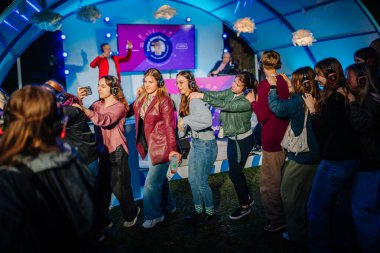 Cesis, Letonya - 20 Haziran 2025 - Sessiz bir disko etkinliğinde neşeli bir grup genç dans ediyor, hepsi kablosuz kulaklık takıyor ve canlı ışıklar altında gülümsüyor..