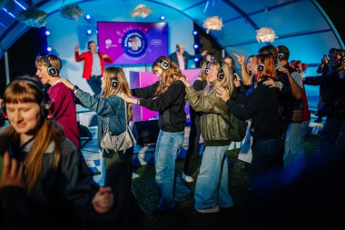 Cesis, Letonya - 20 Haziran 2025 - Sessiz bir disko etkinliğinde canlı bir konga kuyruğunda dans eden gençler, hepsi kulaklık takıyor ve renkli sahne ışıkları altında müziğin tadını çıkarıyorlar..