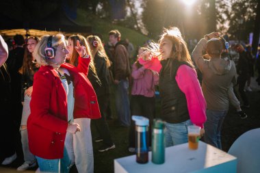 Cesis, Letonya - 20 Haziran 2025 - İki kadın gün batımında, açık havada, sessiz bir disko etkinliği sırasında dans ediyor ve gülümsüyor, parlak kulaklıklar takıyor ve neşeli bir kalabalıkla çevrili müzik dinliyorlar..