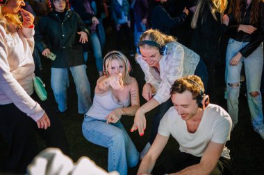 Cesis, Letonya - 20 Haziran 2025 - Genç yetişkinler hareketli bir gece atmosferinde renkli kablosuz kulaklıklar takarak, açık havada sessiz bir diskoda enerjik bir şekilde dans edip etkileşime geçerler..