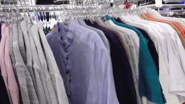 iera, Letonya - 8 Temmuz 2025 - Parlak mağaza ışıklandırması altında seçenekleri gözden geçiren bir kişi ile perakende giyim mağazasında askılar üzerinde düzgün düzenlenmiş gömlekler ve pololar.