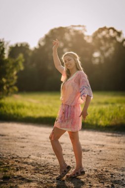 Renkli bir boho tarzı elbise içindeki neşeli kadın ve sandaletler açık havada, yumuşak akşam güneşinde serbestçe dans ediyor. Kaygısız yaz enerjisi yayıyor..