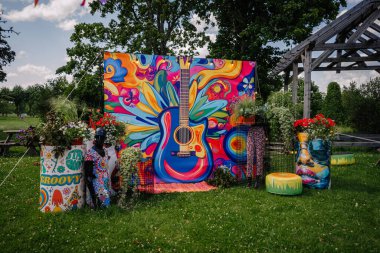 Sigulda, Letonya - 18 Temmuz 2025 - Canlı hippi temalı açık hava dekorasyonu geniş renkli gitar arkaplanı, manken, bitkiler ve fıçılar ve yastıklar üzerine psikedelik sanat eserleri.