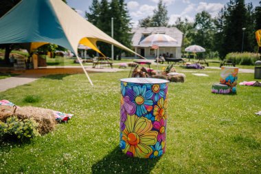 Sigulda, Letonya - 18 Temmuz 2025 - Güneşli bir yaz gününde piknik bankları, şemsiyeler ve dekoratif çadırlarla dolu canlı bir açık hava ortamında renkli çiçek boyalı fıçı.