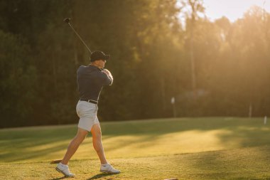 Erkek golfçü, verimli bir golf sahasında, ağaçların yakınındaki engebeli araziden topa vuruyor. Çerçevenin yanında golf çantalı bir itme arabası duruyor..