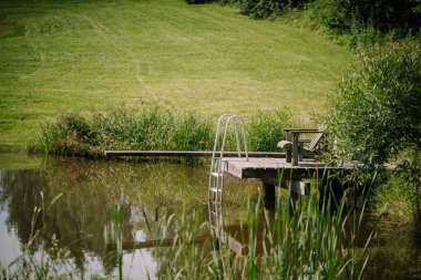Yeşil çimenler ve kamışlarla çevrili küçük ahşap bir rıhtım ve bir sandalyenin olduğu huzurlu bir yaz sahnesi. Rahatlamak ve yüzmek için mükemmel..