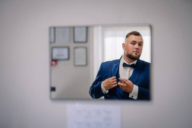 Mavi takım elbiseli ve papyonlu bir damat, aynadan kendine bakarken ceketini düzeltir ve resmi veya düğün etkinliğine hazırlanır..