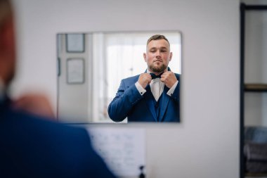 Mavi takım elbiseli bir damat papyonunu düzeltirken aynaya güvenle bakar ve modern, aydınlık bir odada düğün gününe hazırlanır..