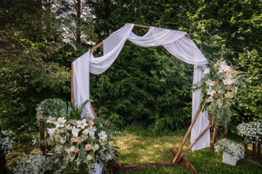 Beyaz kumaş ve çiçeklerle süslenmiş güzel bir açık hava düğün kemeri. Beyaz zambaklar, şeftali gülleri ve orman düzeninde yeşillik..