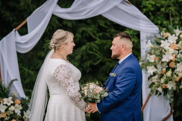 Gelin ve damat, açık hava düğün törenlerinde, beyaz kumaşla süslenmiş bir çiçek kemerinin altında el ele tutuşup birbirlerine bakıyorlar..