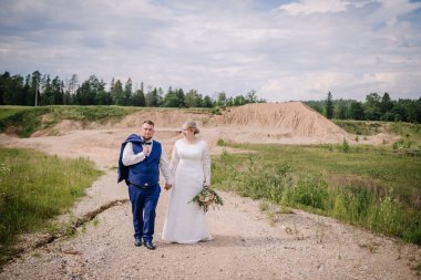Gelin ve damat, doğa ve kumlu tepelerle çevrili çakıllı bir yolda el ele yürür. Damat ceketini omuzunda taşır ve gelin bir buket çiçek tutar..