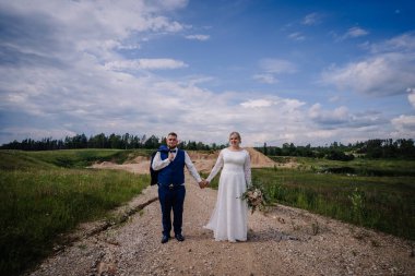 Gelin ve damat, doğa ve kumlu tepelerle çevrili çakıllı bir yolda el ele yürür. Damat ceketini omuzunda taşır ve gelin bir buket çiçek tutar..