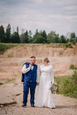 Gelin ve damat kırsal alanda el ele yürüyorlar. Damat, birbirlerine gülümserken tesadüfen ceketini omuzunda taşır..